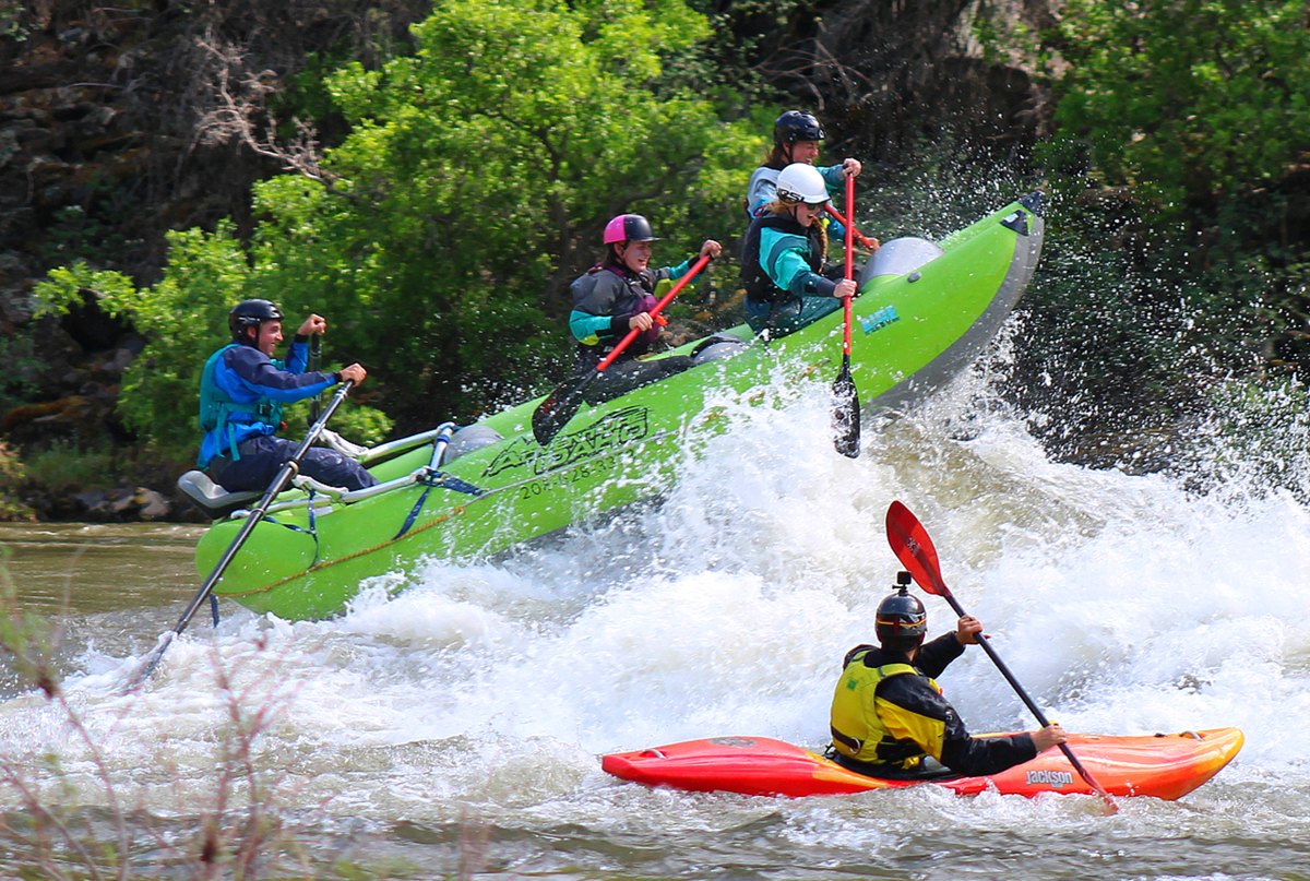 A raft of guides in drysuits and helmets driving through a large breaking wave during preseason training, the boat tipped up on edge, with a safety kayaker holding position in the foam below.