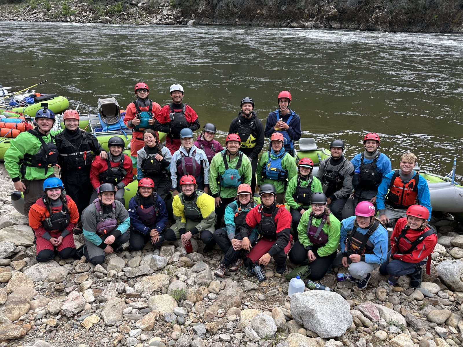 Roughly thirty river guides in drysuits and helmets, gathered on a rocky beach beside rafts on the Salmon River in Idaho.