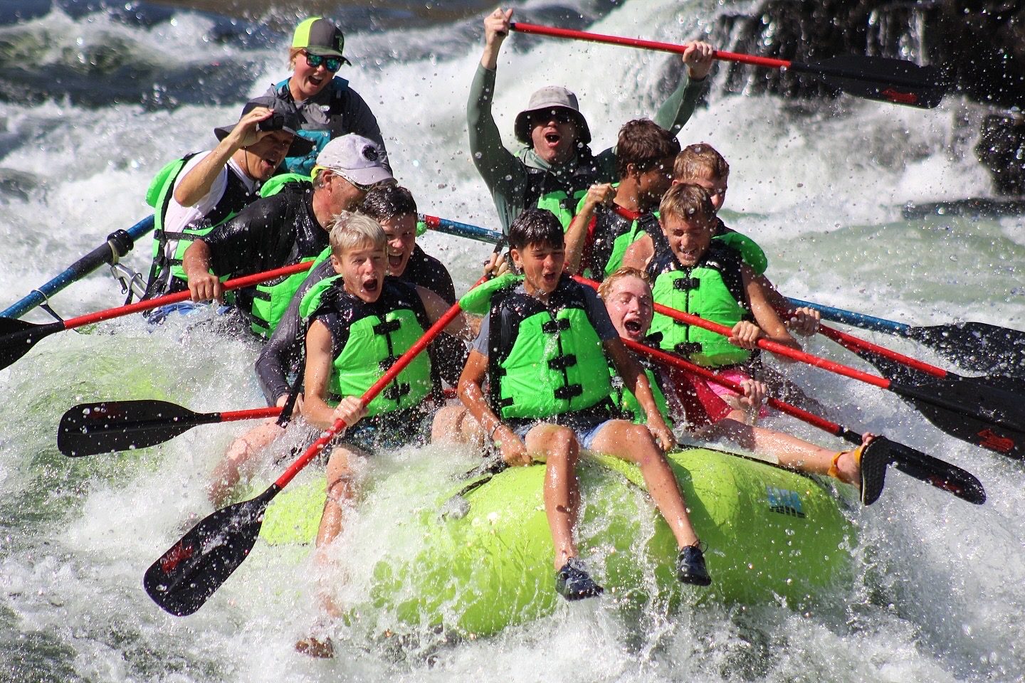 A raft full of teenagers and guides dropping through a whitewater rapid, paddles raised, water breaking over the bow.